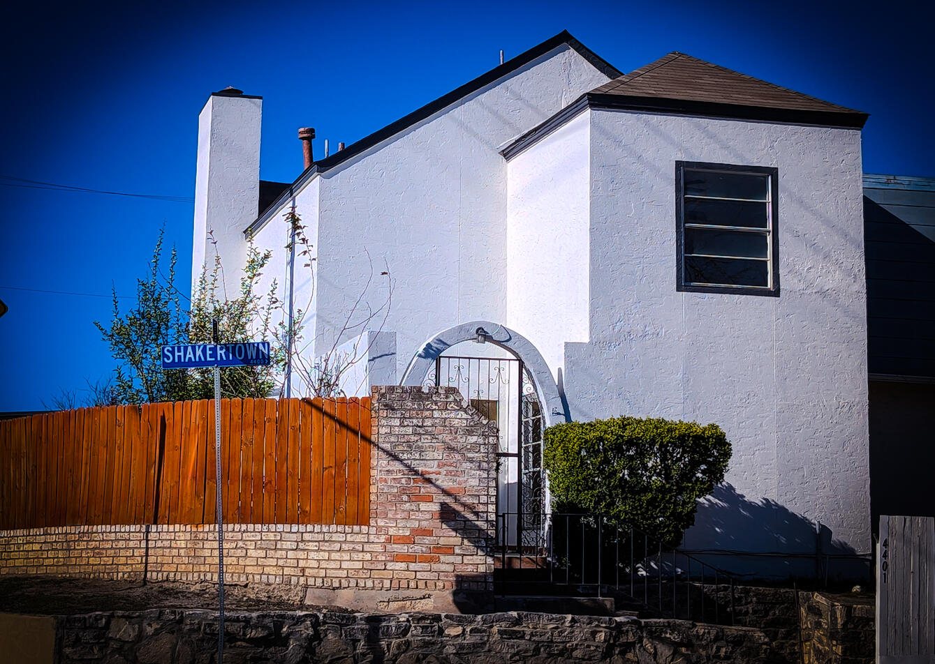 Front of House can be seen, white walls, and white archway with a small staircase and bushes lead to the entry. You can also see the brick and wood fence and the street sign reading Shakertown