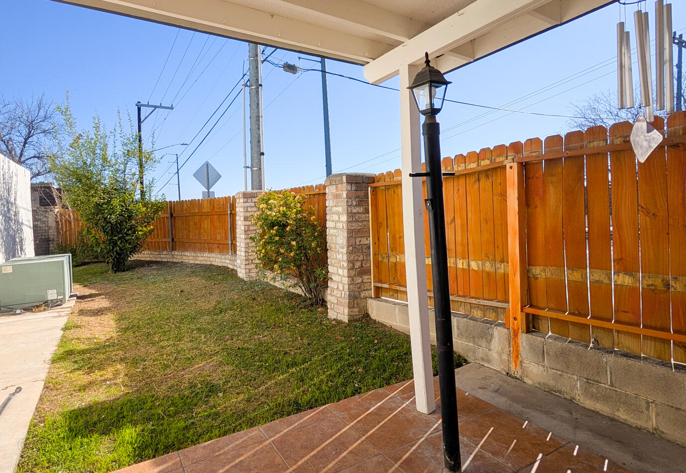 The tiled patio has a light sensor lantern and the covered patio area extends to the fence to the front, and to the right ends a few feet away leading to a grassy section of the yard with a fruit tree and rose bush