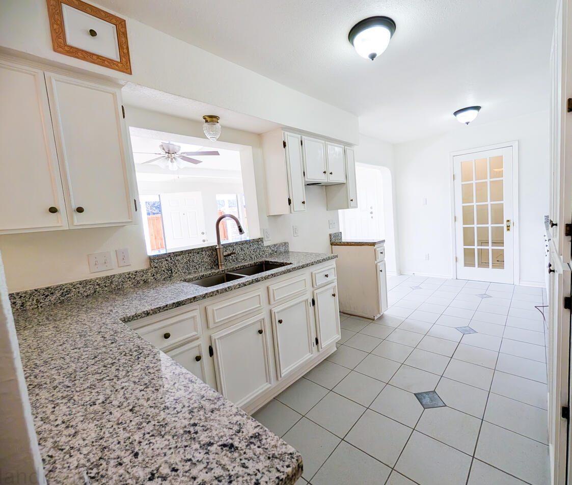 White walls and cabinets can be seen, as well as a large two section stainless sink with brushed nickel faucet and Granite countertops. A light blue floor leads to the Laundry area with a Vintage glass paneled wooden door