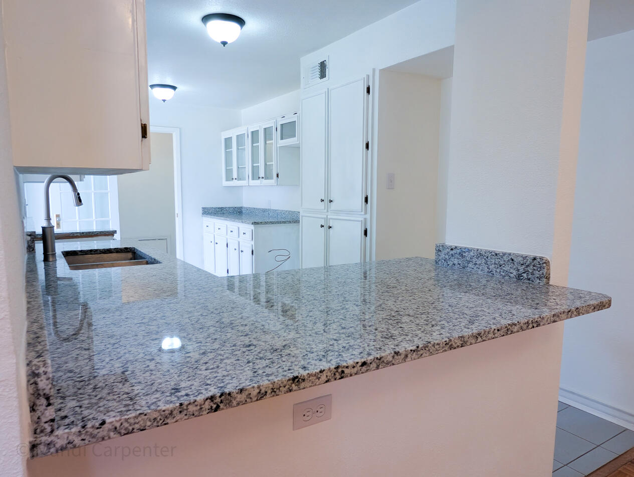 White walls and a solid and warmly flecked granite counter are seen along with a wall of cabinets and the space for fridge as well as a butlers pantry area and separate granite topped bar